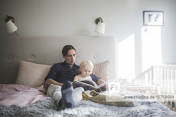 Vater liest mit Kleinkind Sohn Bilderbuch  während er zu Hause auf dem Bett sitzt