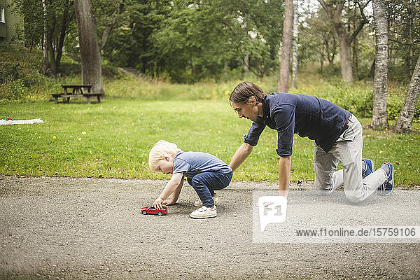 Vater kniet  während der Sohn mit einem Spielzeugauto auf dem Fußweg gegen Pflanzen im Park spielt