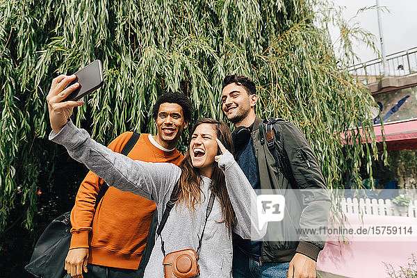 Lächelnde Freunde beim Selbermachen mit dem Handy  während sie in der Stadt am Baum stehen