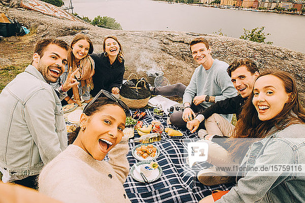 Porträt von fröhlichen Freunden  die im Sommer auf dem Feld am See picknicken