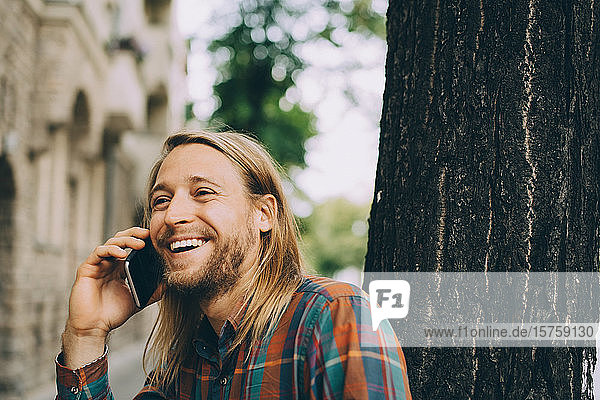 Smiling man talking on mobile phone while leaning on tree trunk in city