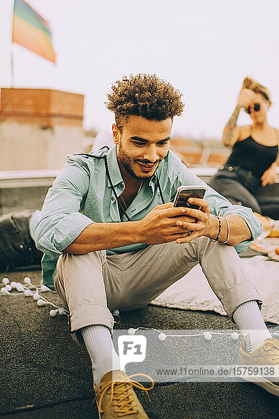 Lächelnder junger Mann benutzt Smartphone  während er bei einer Party auf der Terrasse sitzt