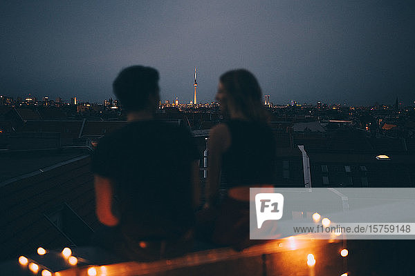 Rückansicht eines Paares  das in der Abenddämmerung auf einer Terrasse gegen das Stadtbild sitzt