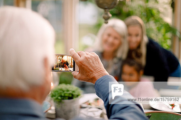 Älterer Mann fotografiert glückliche Familie mit Mobiltelefon während des Mittagessens