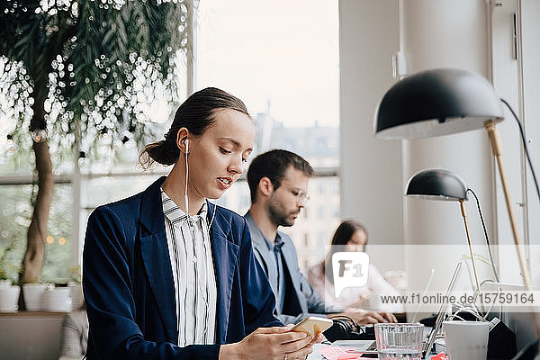 Businesswoman listening music through headphones while sitting with colleagues at desk in office