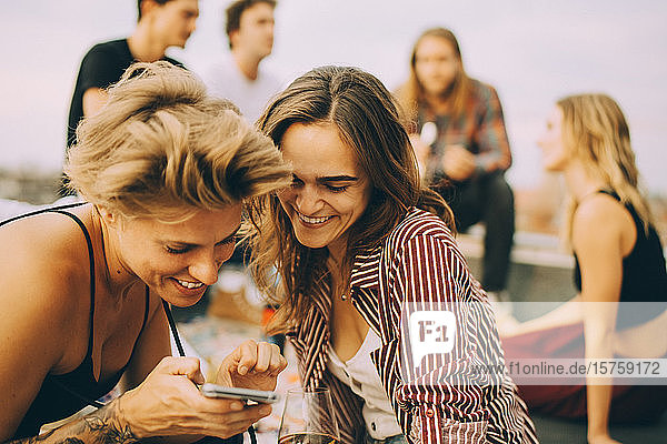Lächelnde Frau zeigt einem Freund ihr Handy  während sich die Leute während der Party auf der Gebäudeterrasse vergnügen