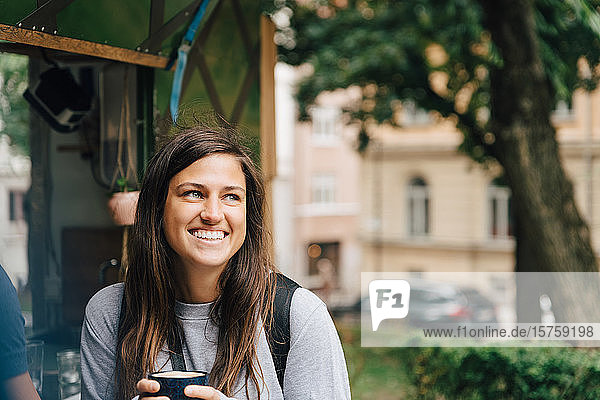 Lächelnde Frau hält Kaffeetasse in der Hand  während sie an einem Imbissstand in der Stadt sitzt