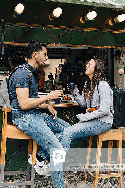 Lächelnde Freunde in voller Länge  die Kaffeetassen in der Hand halten  während sie an einem Imbissstand in der Stadt sitzen