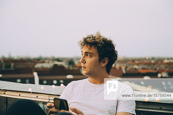 Nachdenklicher Mann schaut weg  während er sein Mobiltelefon auf einer Terrasse in der Stadt gegen den Himmel hält