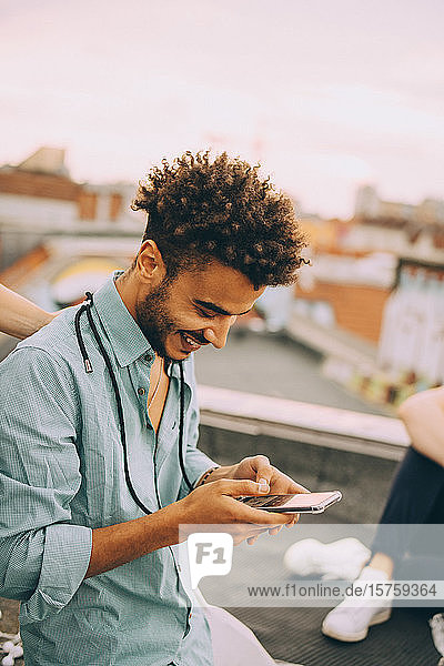 Lächelnder junger Mann per SMS auf Smartphone auf Terrassenparty