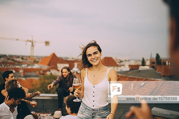 Porträt einer fröhlichen Frau  die Bier genießt  während sie sich mit Freunden auf einer Dachparty amüsiert
