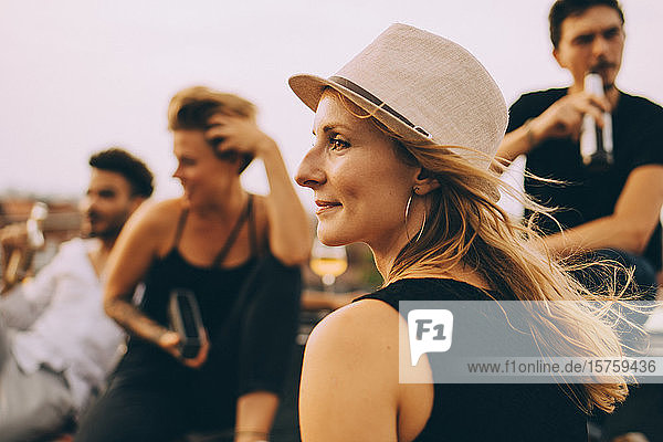 Nachdenkliche junge Frau mit Hut schaut weg  während sie mit Freunden auf der Terrasse sitzt