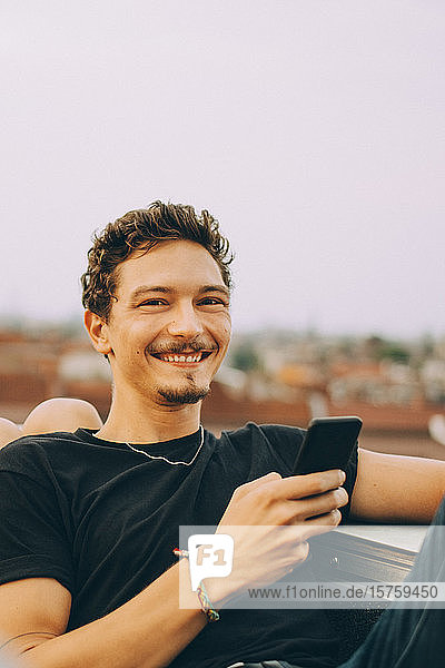 Portrait of smiling young man using mobile phone while sitting on terrace against sky