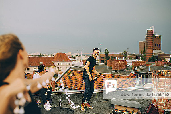 Cheerful man and woman playing with string light on terrace during rooftop party
