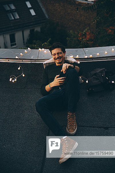 Fröhlicher Mann in voller Länge beim Mobiltelefonieren  während er sich während der Party in der Abenddämmerung auf der beleuchteten Terrasse entspannt