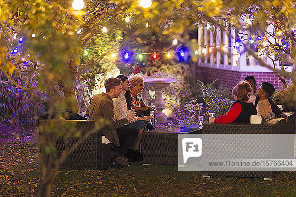 Freunde unterhalten sich und trinken unter Bäumen mit Lichterketten bei einer Gartenparty