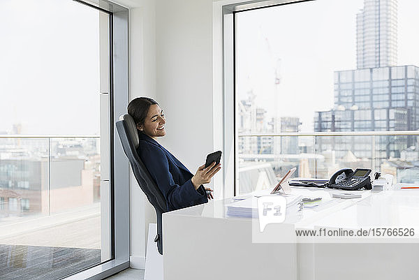 Lächelnde Geschäftsfrau  die in einem städtischen Büro ein Smartphone benutzt