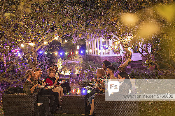 Freunde unterhalten sich und essen Nachtisch unter Bäumen mit Lichterketten bei einer Gartenparty