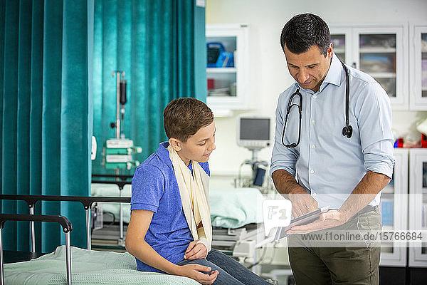 Male pediatrician showing digital tablet to boy patient with arm in sling in hospital