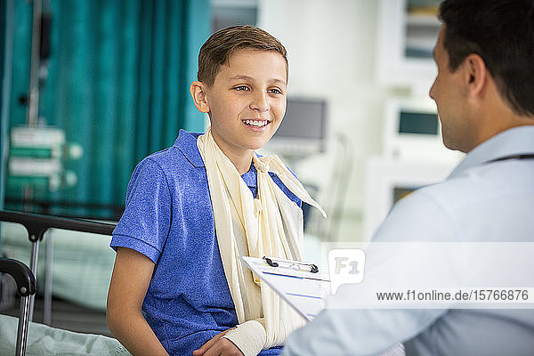 Männlicher Kinderarzt im Gespräch mit einem jungen Patienten mit Arm in einer Schlinge in einer Klinik