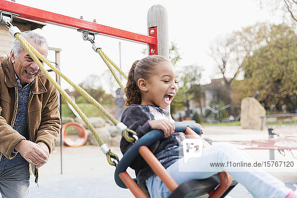 Playful grandfather pushing granddaughter on playground swing