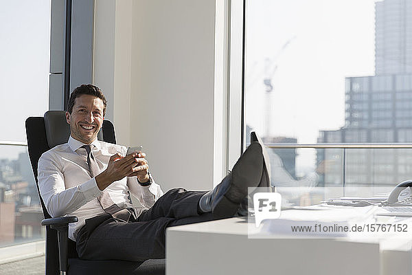 Porträt eines selbstbewussten Geschäftsmannes  der sein Smartphone benutzt und die Füße auf den Schreibtisch in einem Hochhausbüro legt