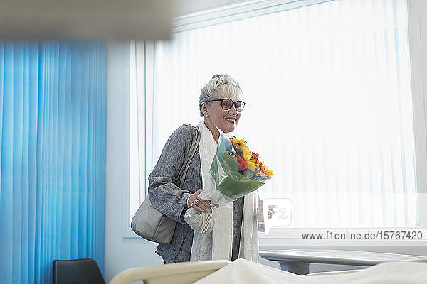 Lächelnde ältere Besucherin mit Blumenstrauß im Krankenhauszimmer