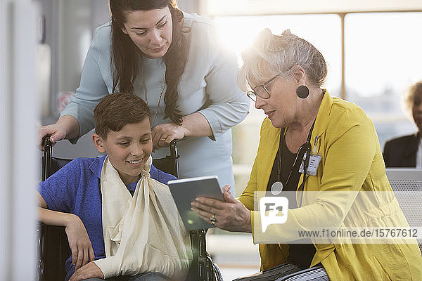 Arzt mit digitalem Tablet im Gespräch mit einem jungen Patienten im Rollstuhl und seiner Mutter in der Kliniklobby