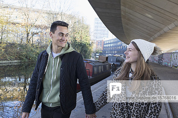 Glückliches junges Paar  das sich an den Händen hält und an einem städtischen Kanal entlanggeht