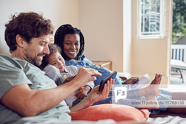 Schwangere junge Familie  die auf dem Sofa ein Smartphone und ein digitales Tablet benutzt