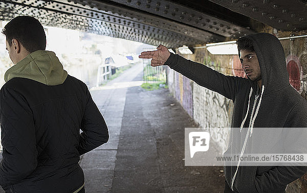 Bedrohlich wirkender junger Mann  der in einem städtischen Tunnel eine Fingerpistole auf einen Mann richtet
