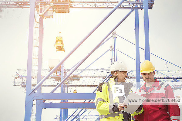 Dock worker and manager with clipboard talking below crane at shipyard
