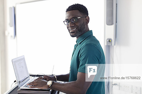 Porträt eines lächelnden  selbstbewussten männlichen Lehrers am Laptop  der sich im Klassenzimmer vorbereitet