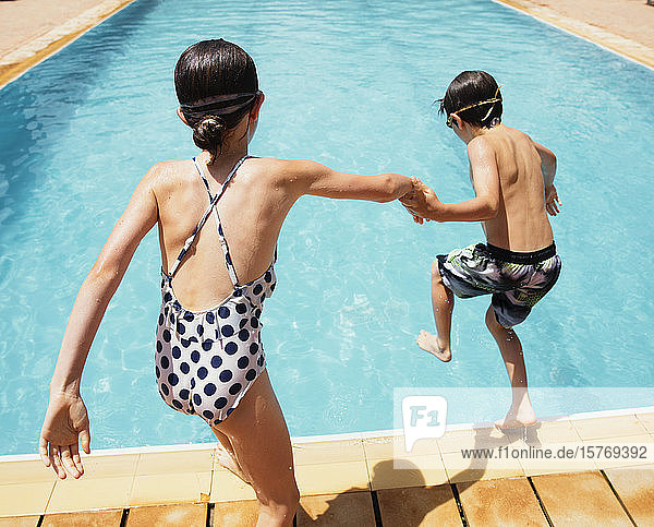 Junge und Mädchen halten sich an den Händen und springen in ein sonniges Sommerschwimmbad