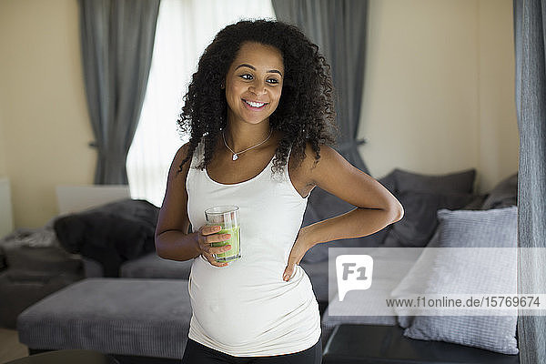 Glückliche junge schwangere Frau trinkt grünen Smoothie im Wohnzimmer