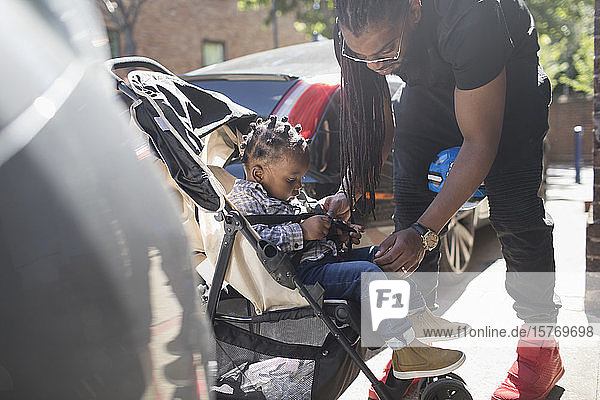 Father fastening toddler son in stroller on sunny sidewalk
