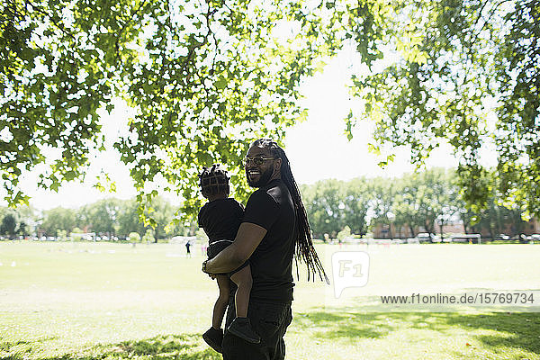 Porträt glücklicher Vater mit langen Zöpfen  der seinen Sohn im sonnigen Park trägt
