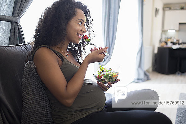 Lächelnde junge schwangere Frau isst Salat auf dem Sofa