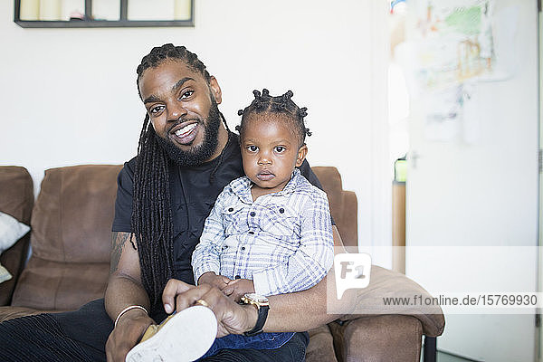 Portrait father with long braids putting shoes on toddler son on sofa