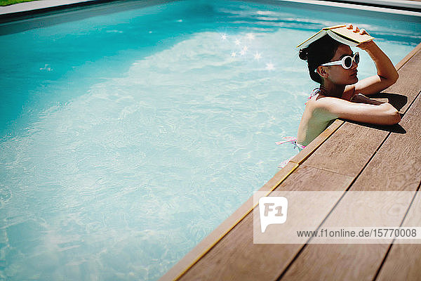 Frau hält Buch über Kopf in sonnigem Sommerschwimmbad