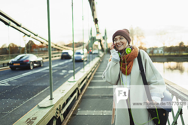 Junge Frau mit Strumpfmütze und Schal spricht auf einer Brücke mit ihrem Smartphone