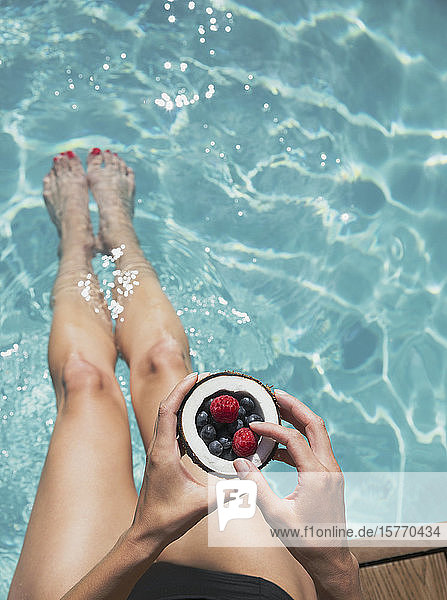 Frau entspannt sich  taucht mit nackten Füßen in den Sommerpool und isst Beeren von der Kokosnuss