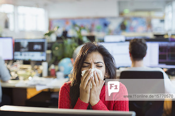 Geschäftsfrau mit Allergien niest am Computer im Büro in ein Taschentuch