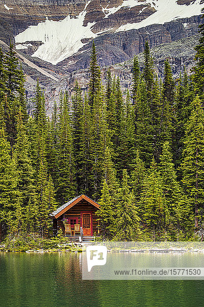 Holzblockhaus an einem Seeufer  das sich in der Sonne spiegelt  mit immergrünen Bäumen und Bergfelsen im Hintergrund; Field  British Columbia  Kanada