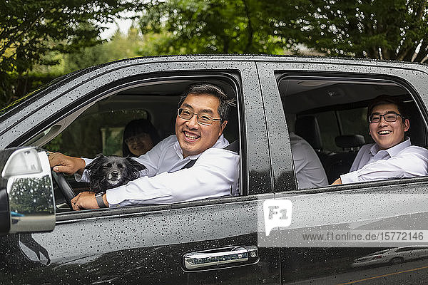 Familienfahrt im Auto mit dem Hund  der Vater am Steuer und die erwachsenen Kinder auf dem Rücksitz  die in die Kamera schauen und lächeln; Langley  British Columbia  Kanada