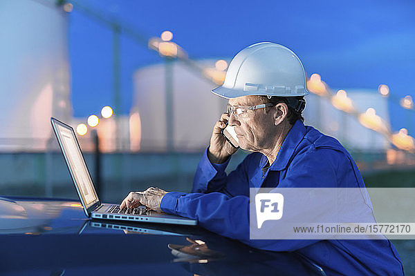 Man working on a laptop and smart phone with an oil refinery in the background; Alberta  Canada