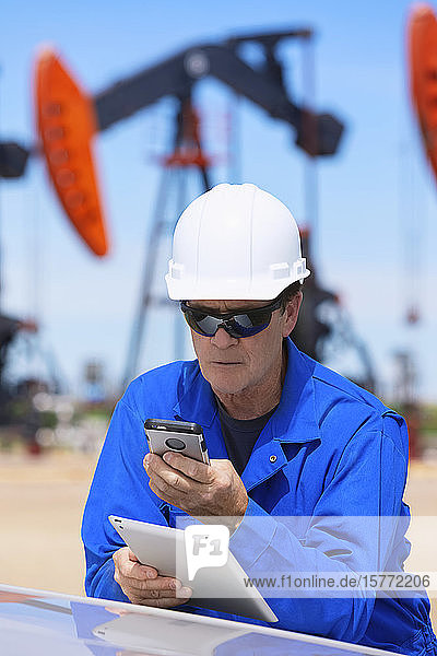Man working on a tablet and smart phone with pumpjacks in the background; Alberta  Canada