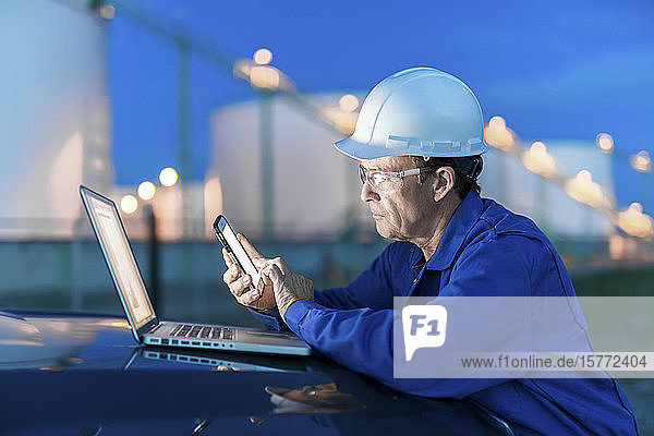 Man working on a laptop and smart phone with an oil refinery in the background; Alberta  Canada