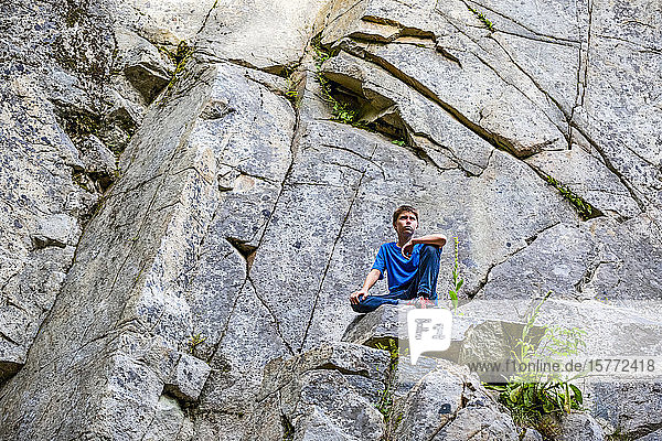 Boy sits on a rock ledge looking out with a serious expression; British Columbia  Canada