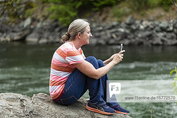 Woman sitting on a rock at the water's edge of Adams River taking pictures with a smart phone; Salmon Arm  British Columbia  Canada